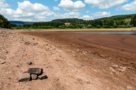 Paisaje de una zona afectada por la sequía, con suelo seco y una pequeña cantidad de agua en el fondo, rodeada de vegetación y montañas